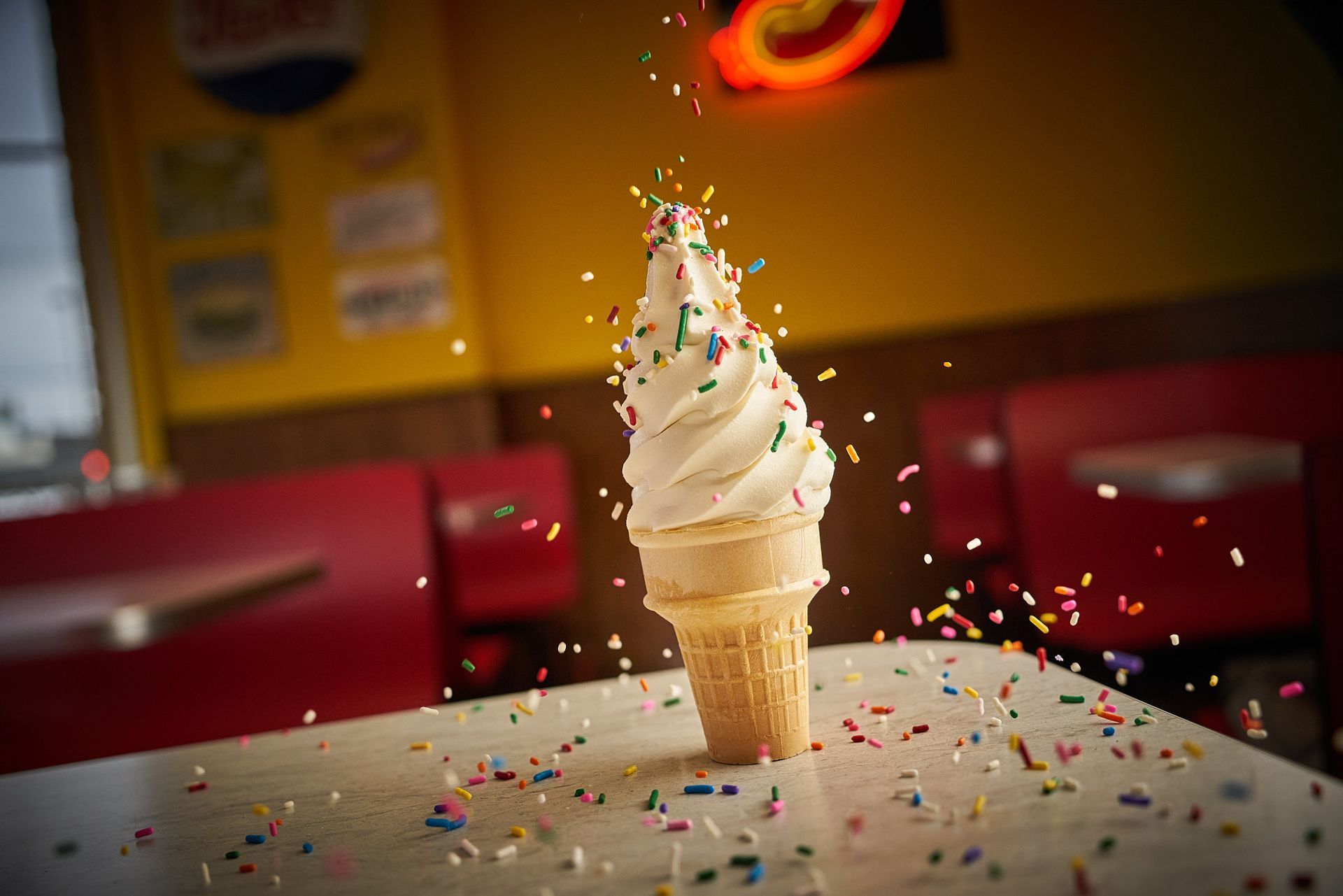 Soft-serve ice cream cone on a table, sprinkles falling; red booth seating and a yellow wall in the background.