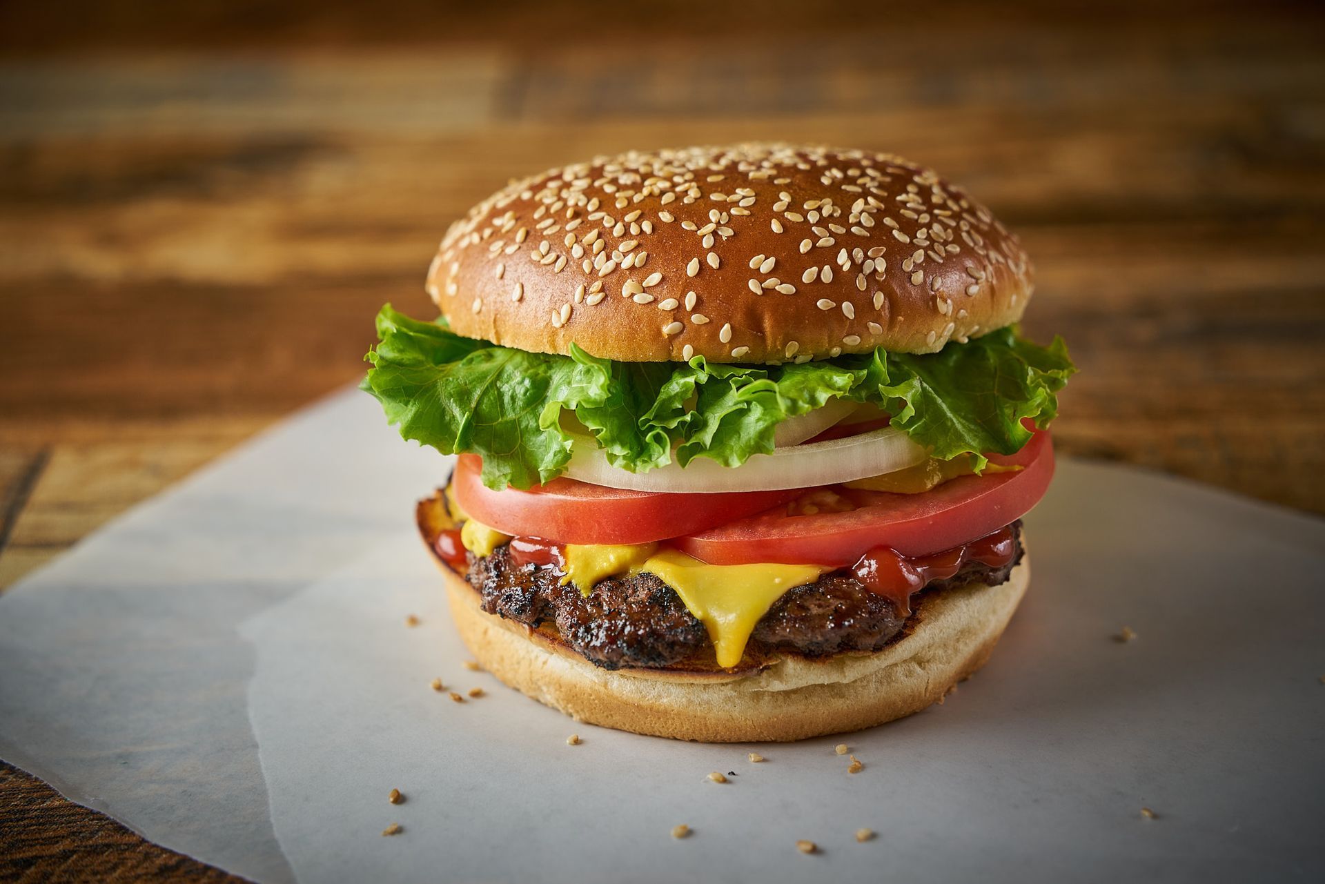 Hamburger with sesame seed bun, lettuce, tomato, onion, cheese, on parchment paper.