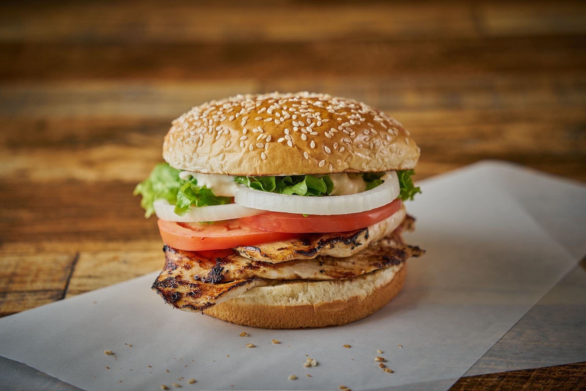 Grilled chicken sandwich with sesame bun, tomato, onion, lettuce. On parchment paper.