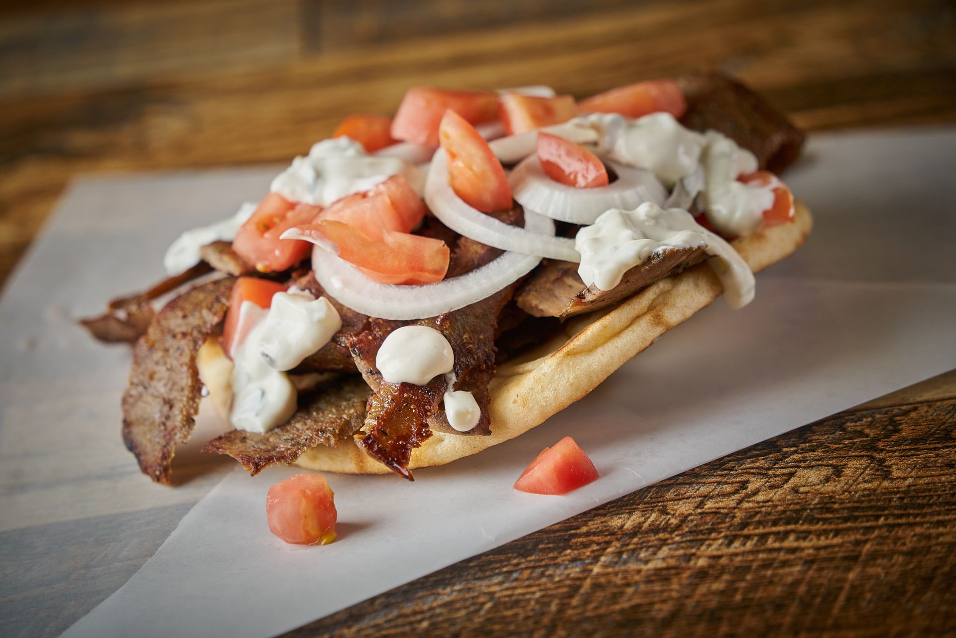 Gyro sandwich on white paper with tomatoes, onions, and white sauce.