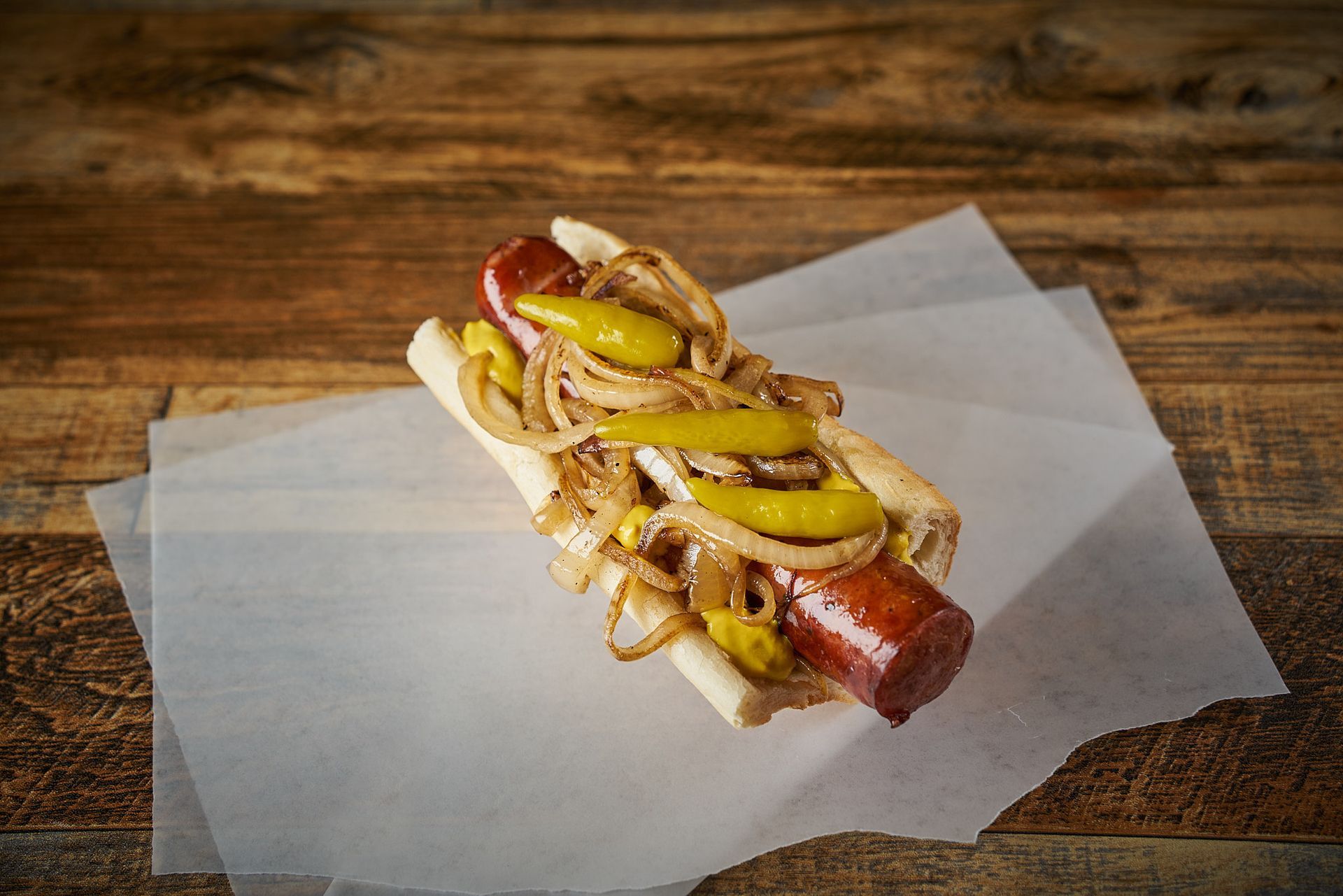 Hot dog on a bun with onions, peppers, and mustard on parchment paper atop wood.