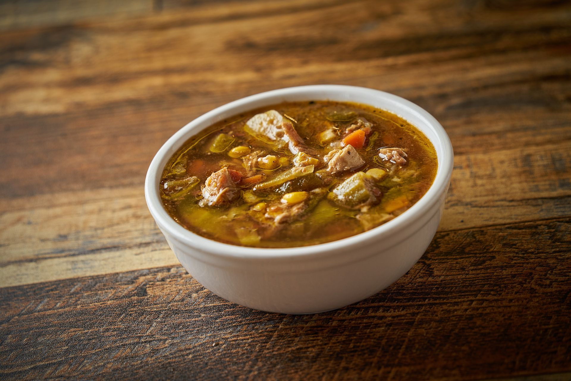 Bowl of gumbo soup on a wooden table, with meat, vegetables, and a dark broth.