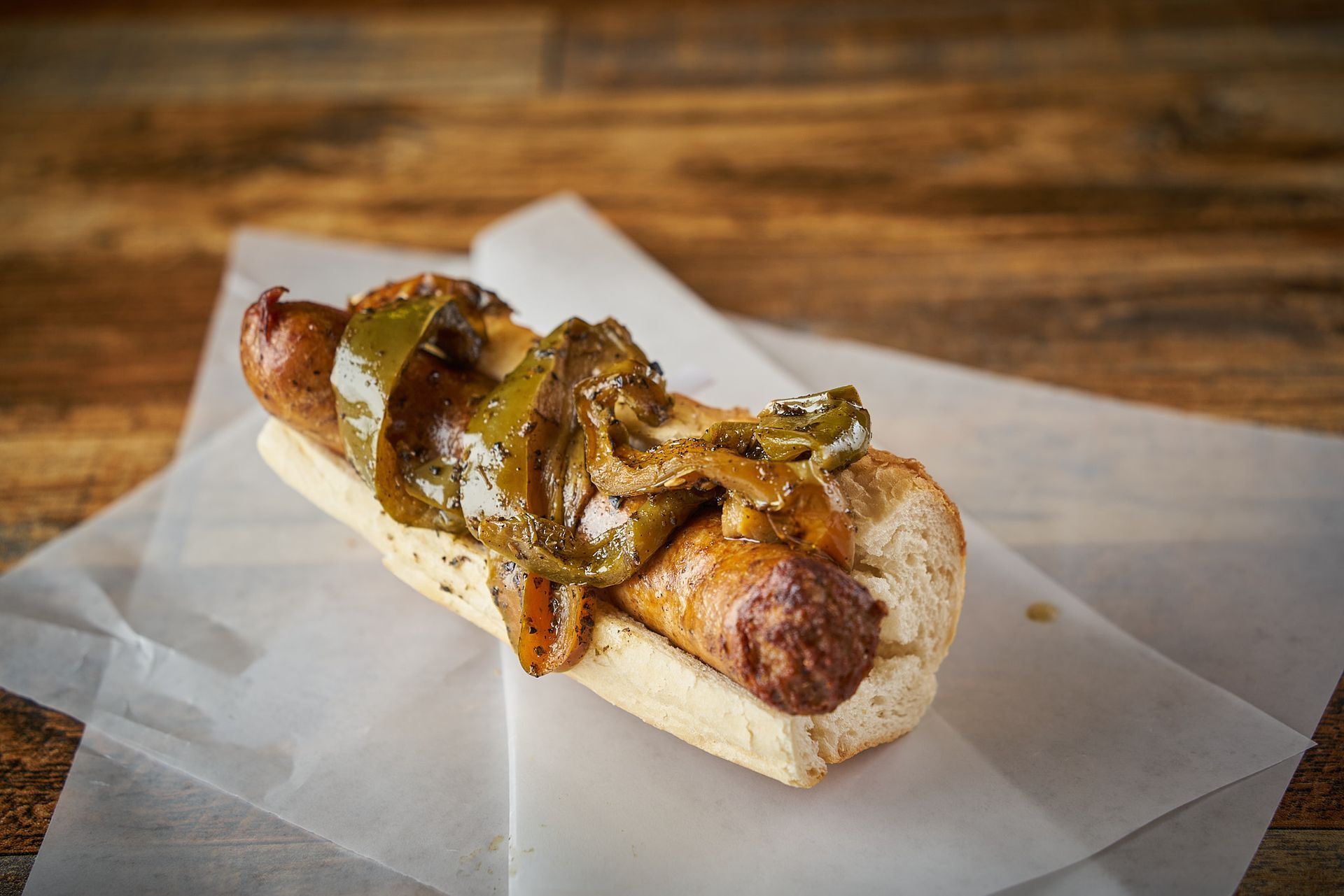 Hot dog with grilled peppers and onions on a white paper, resting on wood surface.