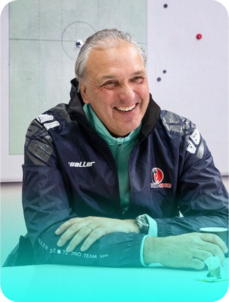 Smiling man in a dark jacket and teal shirt, sitting at a table with a whiteboard in the background.