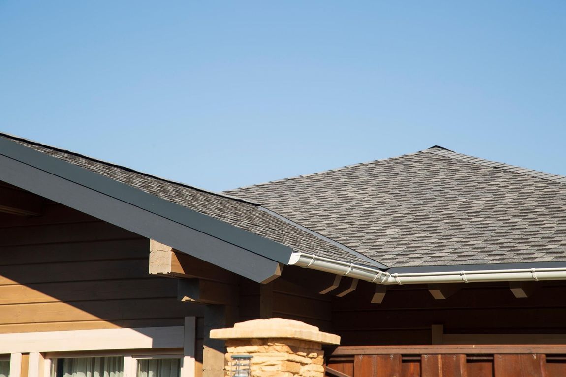 House roof with gray shingles, dark trim, and a stone chimney against a clear blue sky