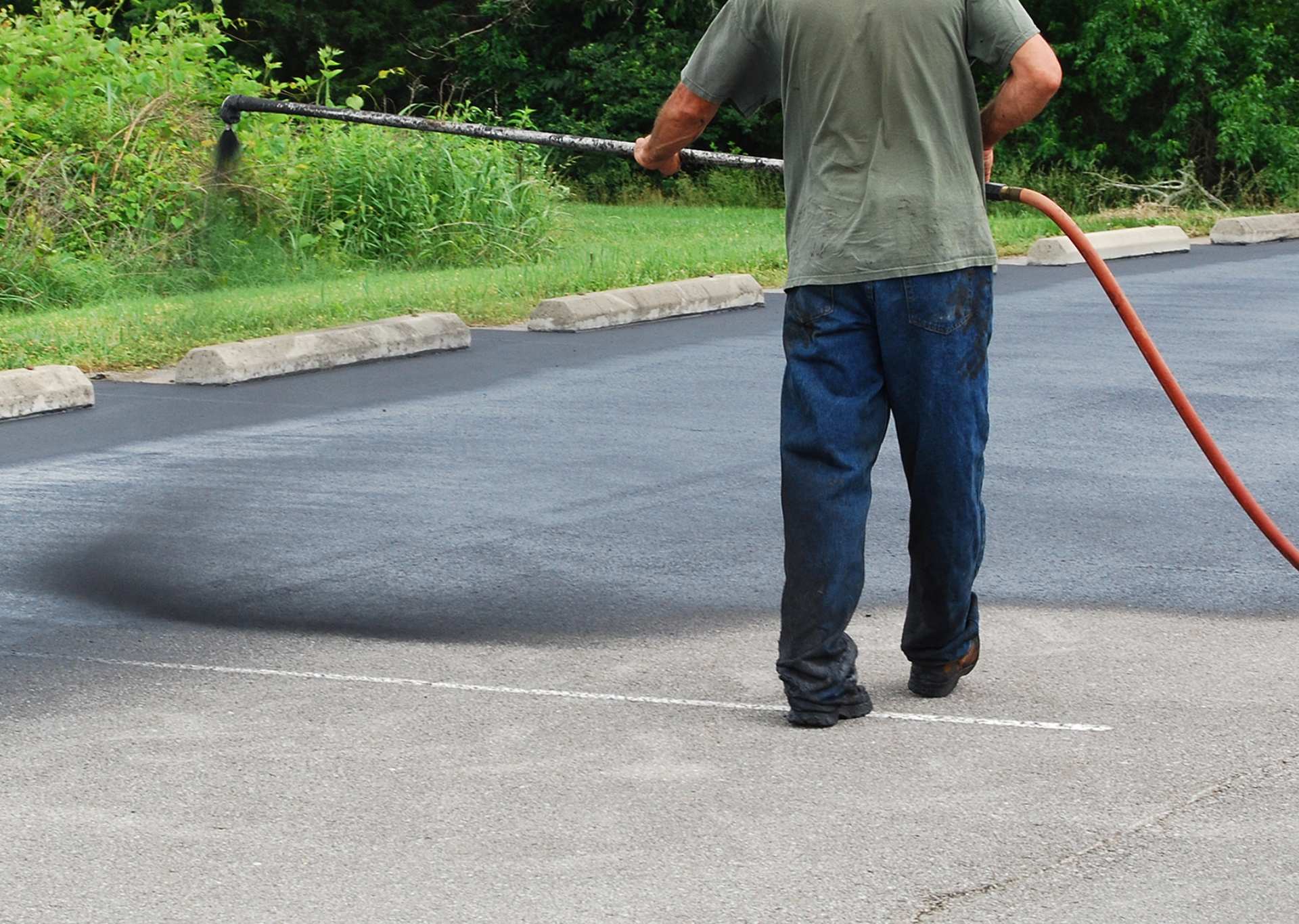 Man applying sealant to a parking lot. He holds a spray wand, applying a black substance.