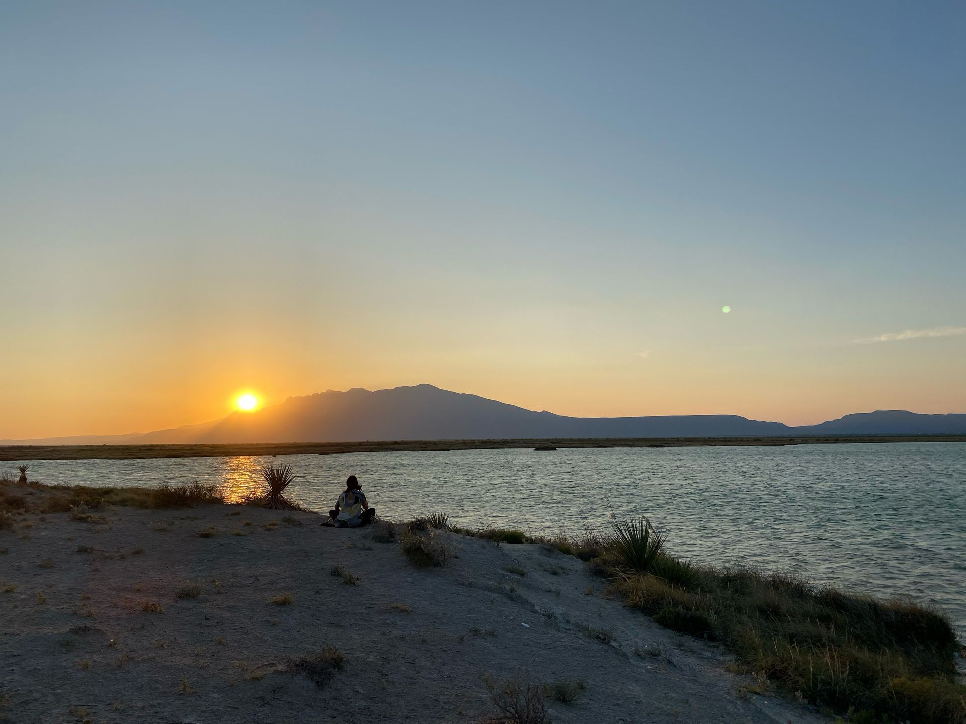 A person is sitting on the shore of a lake at sunset.