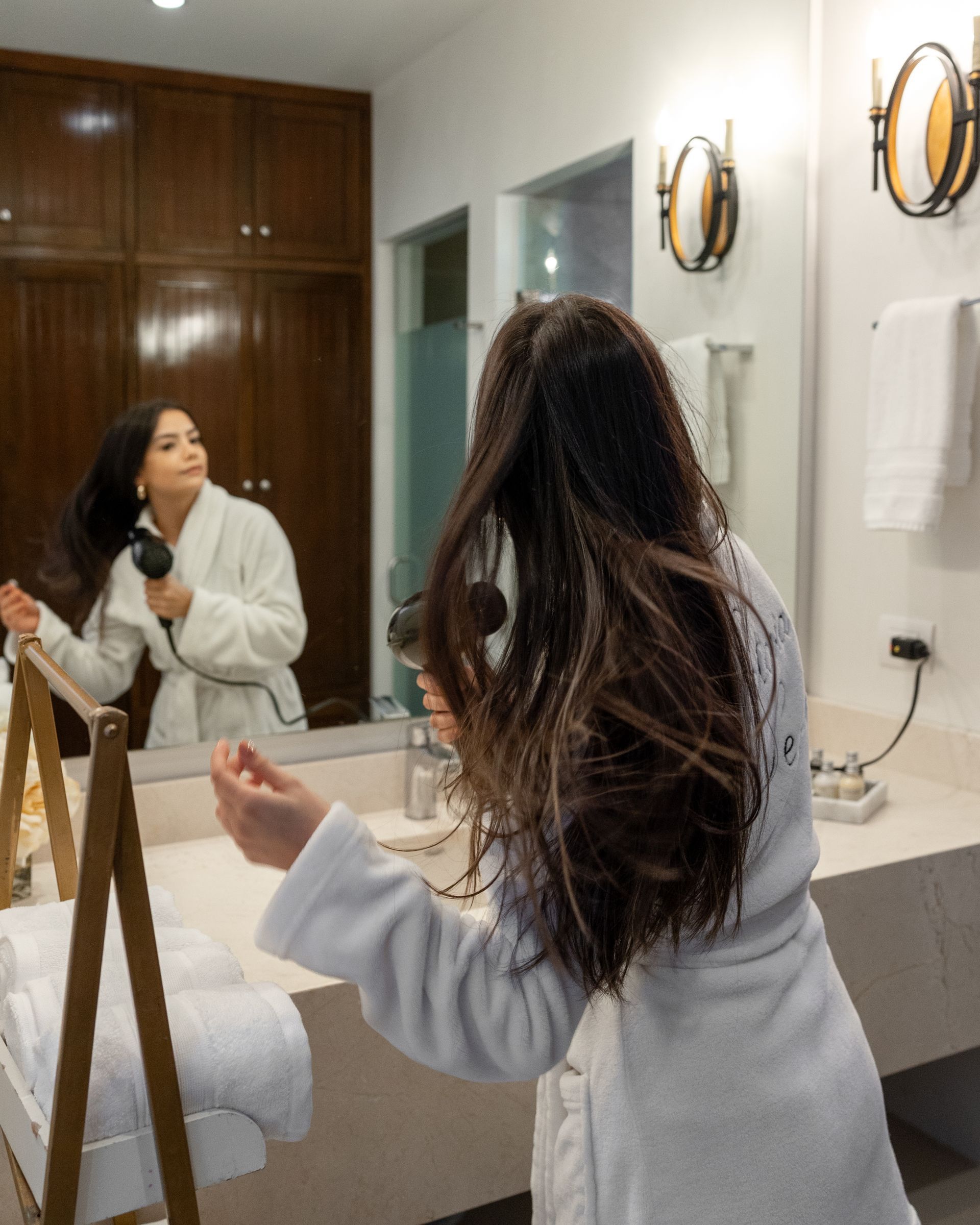 Una mujer en bata se seca el cabello frente a un espejo.