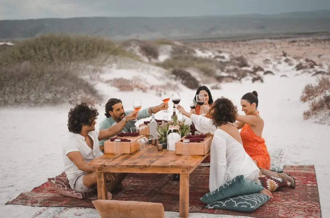 Grupo de amigos brindando con copas de vino en un picnic en la playa. Mesa de madera, alfombra colorida y paisaje árido.