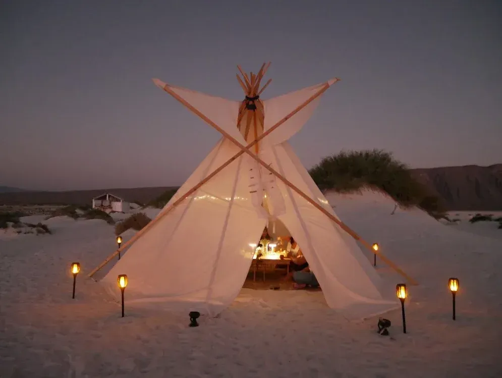 Tipi iluminado en una playa de arena al anochecer, con antorchas tiki que lo rodean.