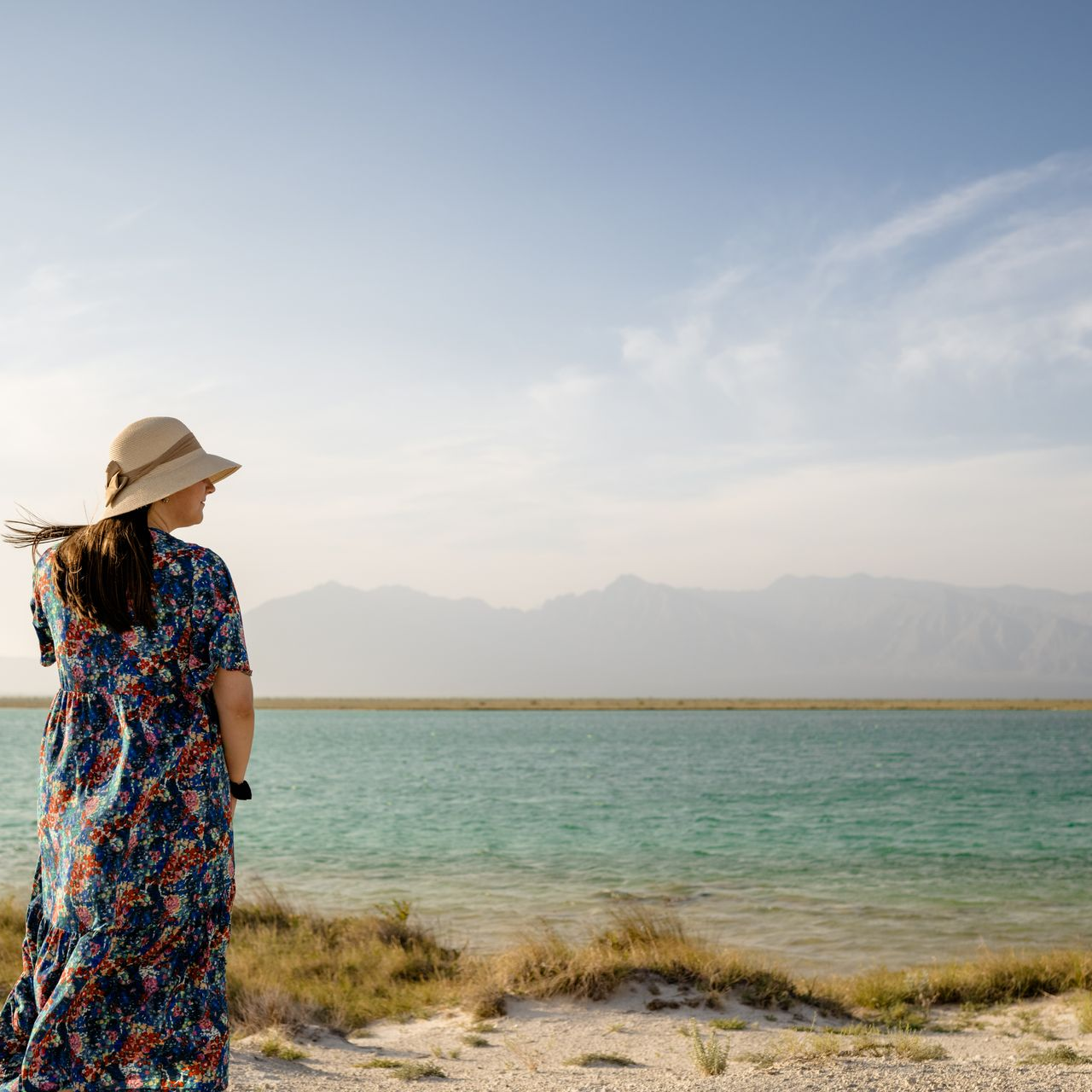 Una mujer con vestido y sombrero está parada en una playa mirando el océano.
