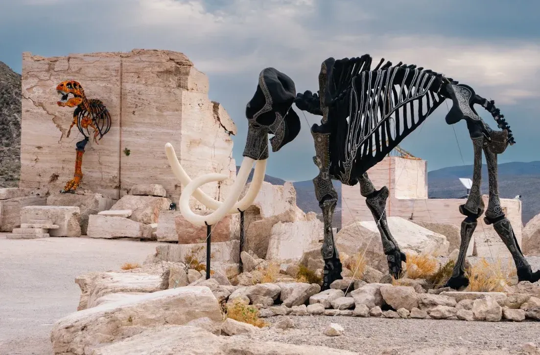 Esqueleto de mamut con colmillos y un esqueleto de pájaro cerca de una estructura de piedra con un fondo de montaña.