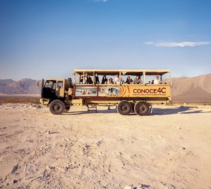 Camión turístico descapotable con pasajeros en un paisaje árido y montañoso, en un día soleado. Logotipo de 
