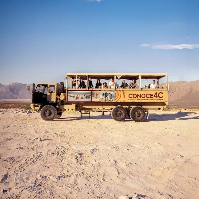 Camión turístico descapotable con pasajeros en un paisaje árido y montañoso, en un día soleado. Logotipo de 