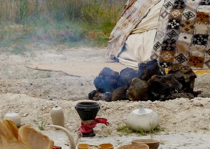 Fogata con humo, objetos en la arena, carpa estampada en el fondo.