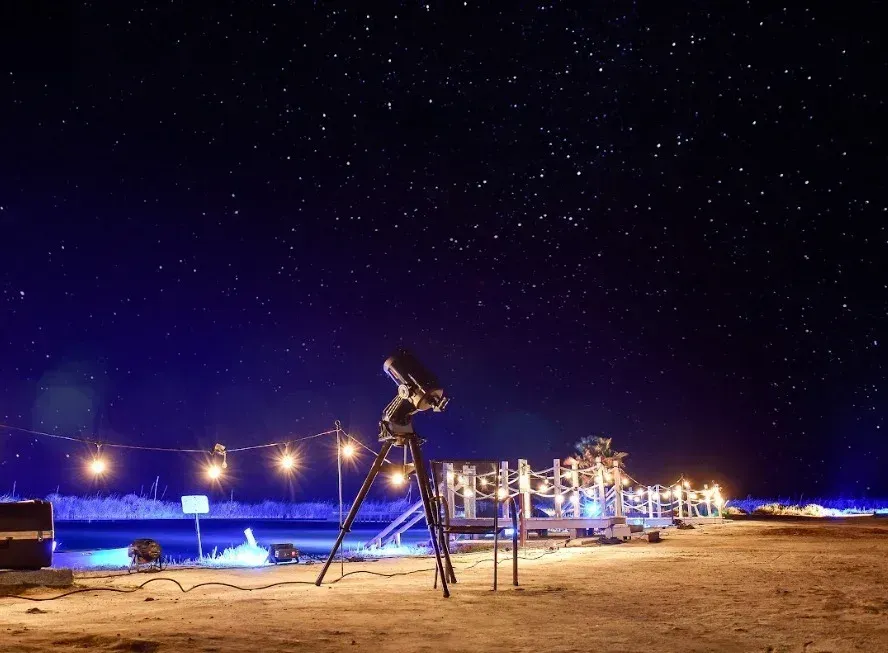 Telescopio apuntando hacia un cielo nocturno estrellado, con luces de cadena iluminando un suelo arenoso y una estructura.