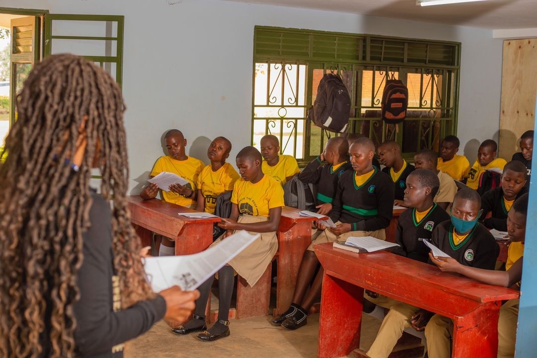 An instructor holds papers while addressing a classroom of students wearing yellow shirts and black uniforms.