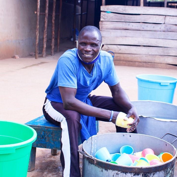 A person in a blue shirt and apron smiles while sitting on a stool, washing colorful cups in a large metal basin.