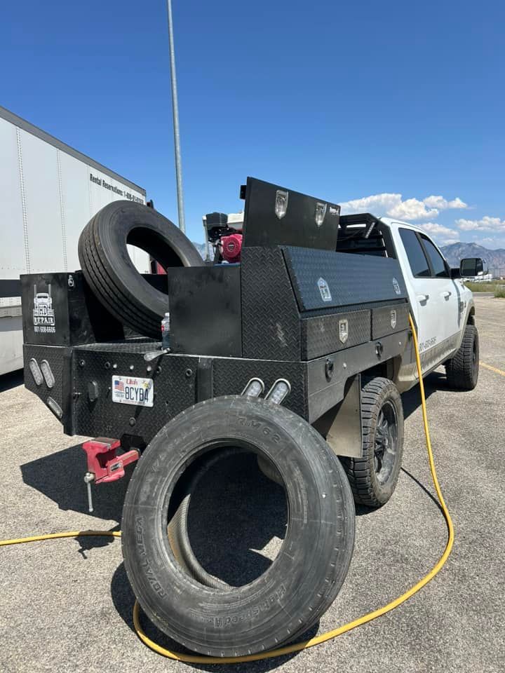 White work truck with a black flatbed, two tires, and toolboxes, parked outdoors on a sunny day.