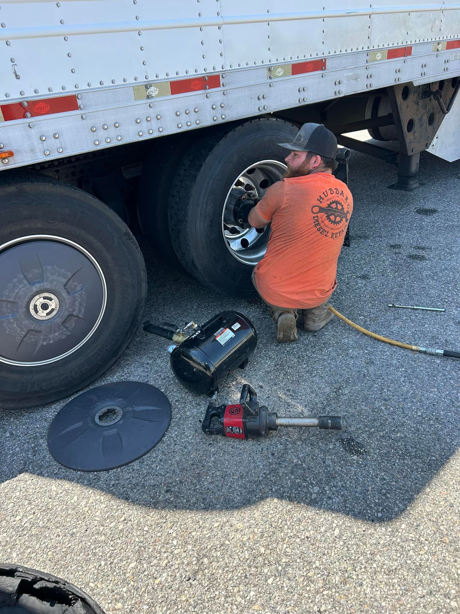 Man in orange shirt kneeling beside a semi-truck, changing a tire with tools on the ground.
