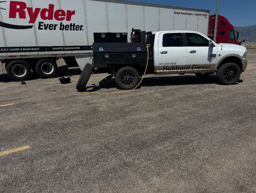 White pickup truck with a black flatbed parked next to a Ryder semi-truck. A spare tire rests on the ground.