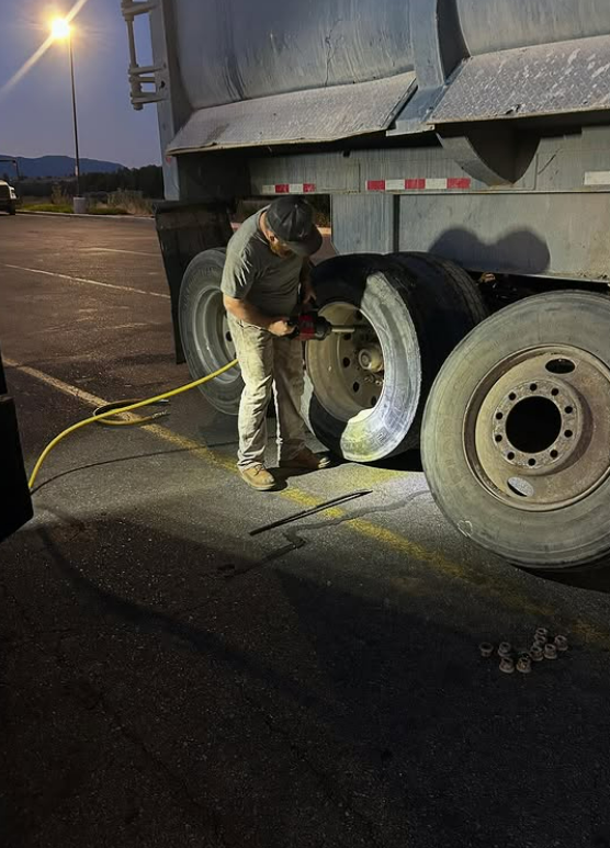 A person works on a semi-truck tire at night, illuminated by a light. They are using a tool connected to a yellow hose.