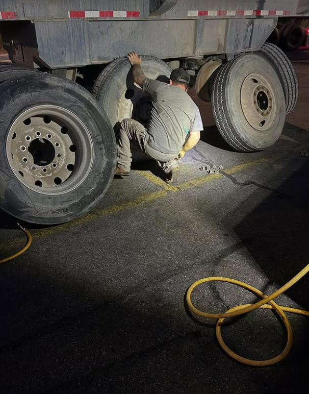 A person is crouched near a semi-truck tire, likely inspecting or repairing it. The scene is on an asphalt surface with yellow lines, possibly a parking area.