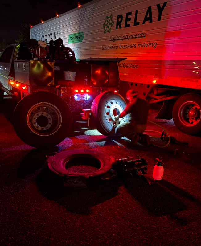 A semi-truck with a flat tire being repaired at night. A person works on the wheel with tools under red emergency lights.