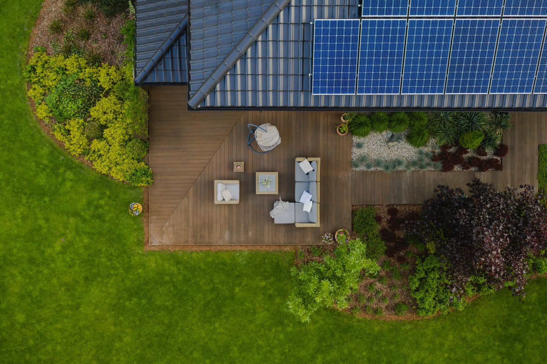 Overhead view of a wooden patio beside a house with outdoor furniture, surrounded by green lawn and shrubs