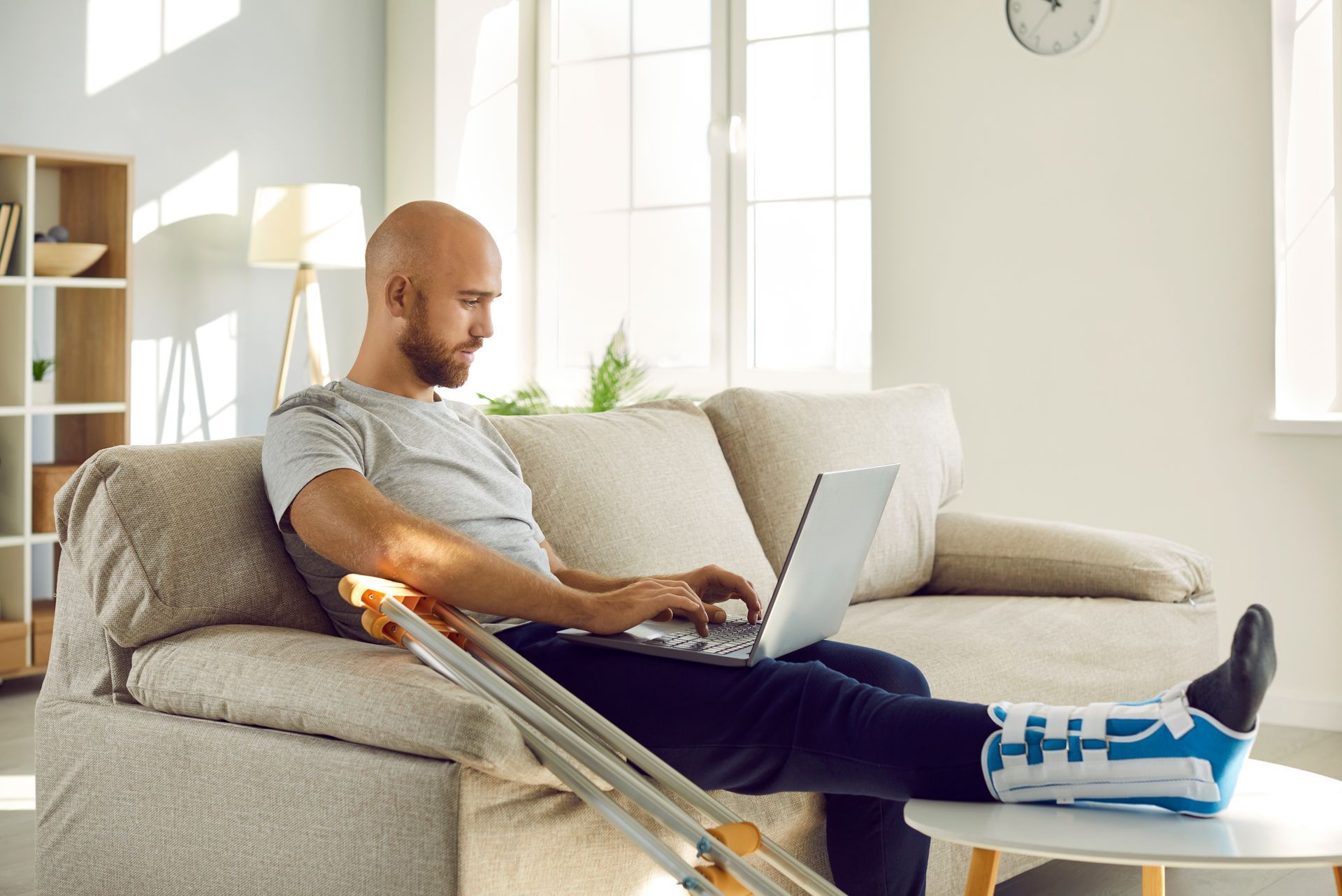 Bald man with leg in cast, working on laptop while sitting on sofa with crutches nearby.