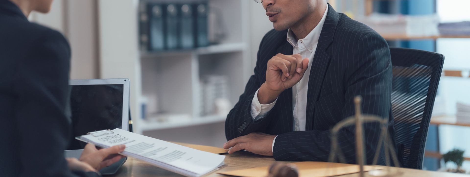 Two professionals in formal attire sit at a desk in an office, reviewing a legal document next to a scale of justice.