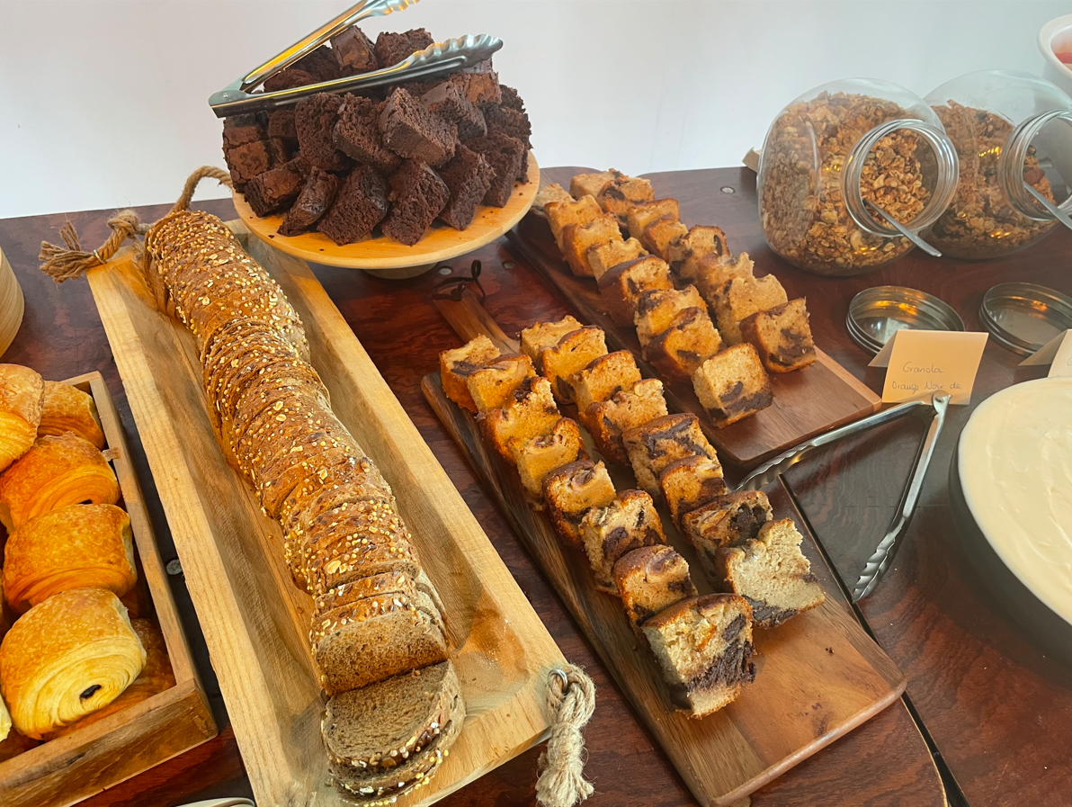 Buffet petit-déjeuner : viennoiseries assorties, pain tranché, brownies, granola et muffins sur une table en bois.