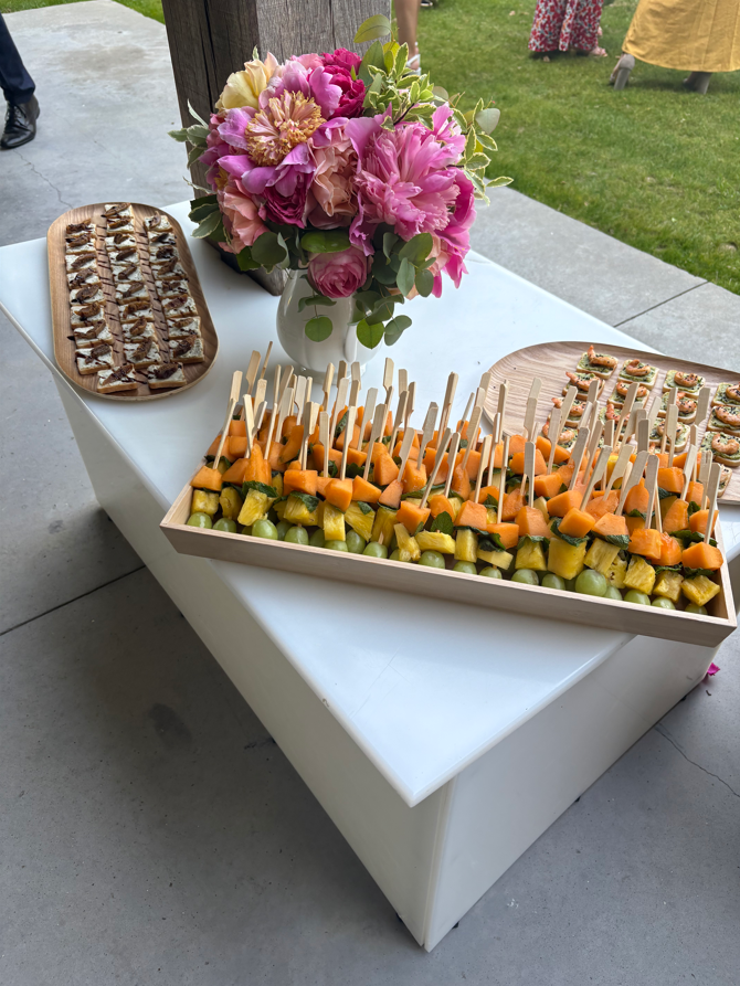 Brochettes de fruits et autres amuse-gueules sur une table blanche, à côté d'un bouquet de fleurs roses.