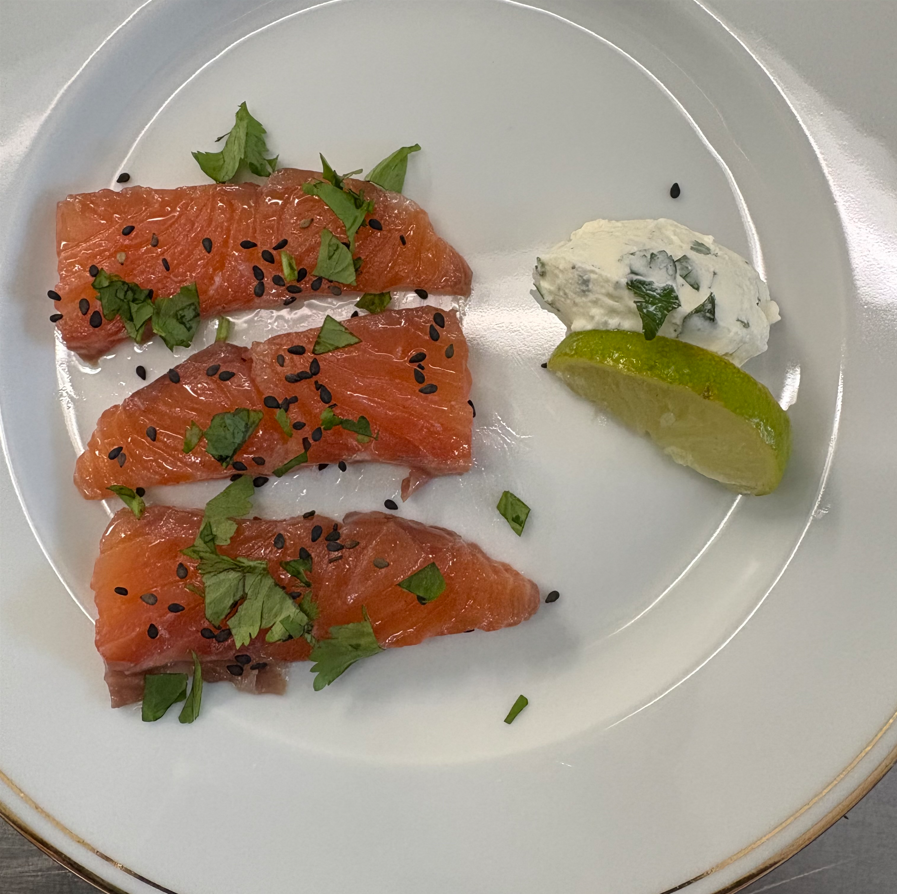 Trois tranches de saumon, beurre aux herbes, quartier de citron vert et graines de sésame noires sur une assiette blanche.