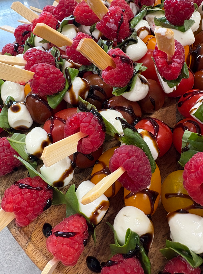 Brochettes de framboises, tomates, mozzarella, basilic avec glaçage balsamique sur planche de bois.