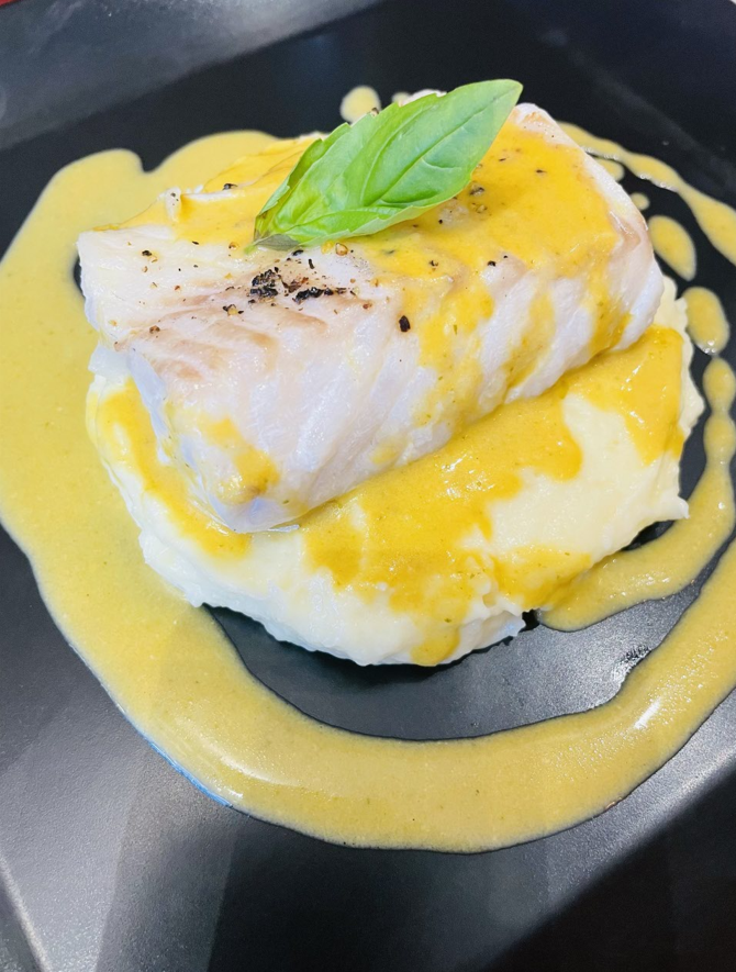 Filet de poisson à la sauce jaune et basilic sur purée de pommes de terre, sur une assiette noire.
