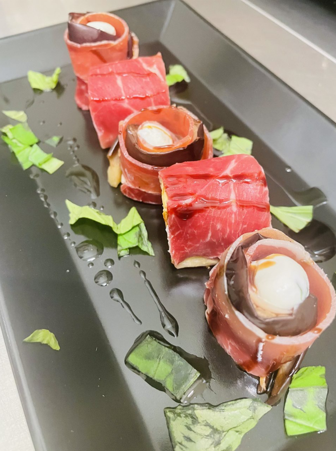 Apéritifs enveloppés de prosciutto sur une assiette noire, arrosés de glaçage balsamique et garnis de basilic.