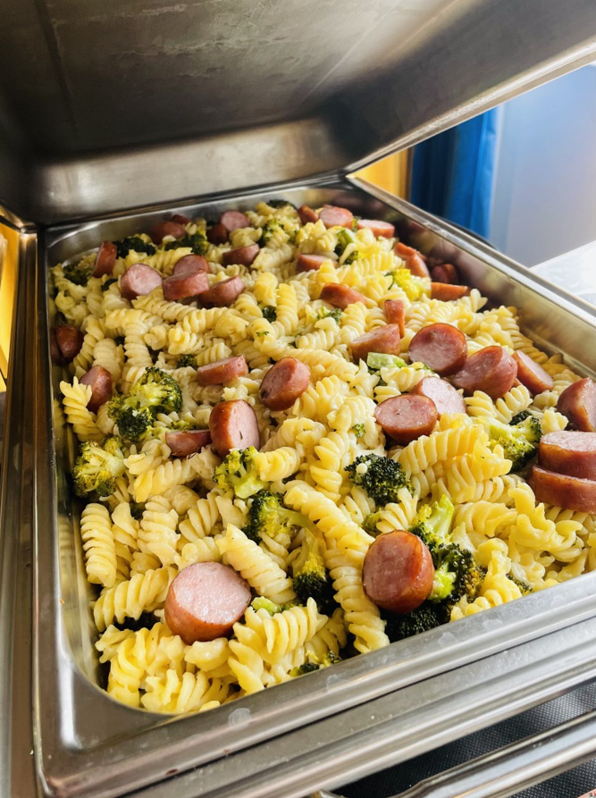 Salade de pâtes avec brocoli et saucisse tranchée dans un chauffe-plats en acier inoxydable.