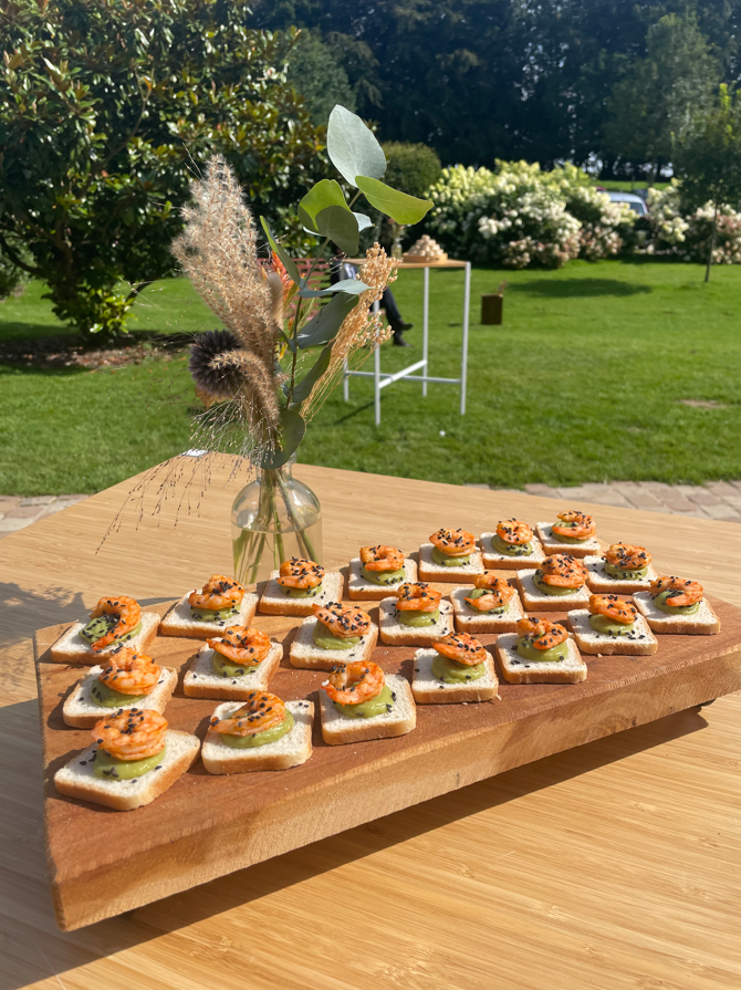 Apéritifs sur toast, disposés sur une planche de bois à l'extérieur avec un vase de fleurs, dans un jardin ensoleillé.