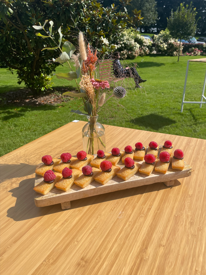 Planche de bois avec des pâtisseries garnies de framboises; vase de fleurs sur une table à l'extérieur dans un jardin.