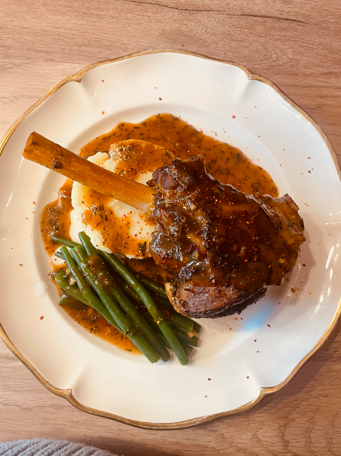Jarret d'agneau avec purée de pommes de terre et haricots verts sur une assiette blanche avec sauce.