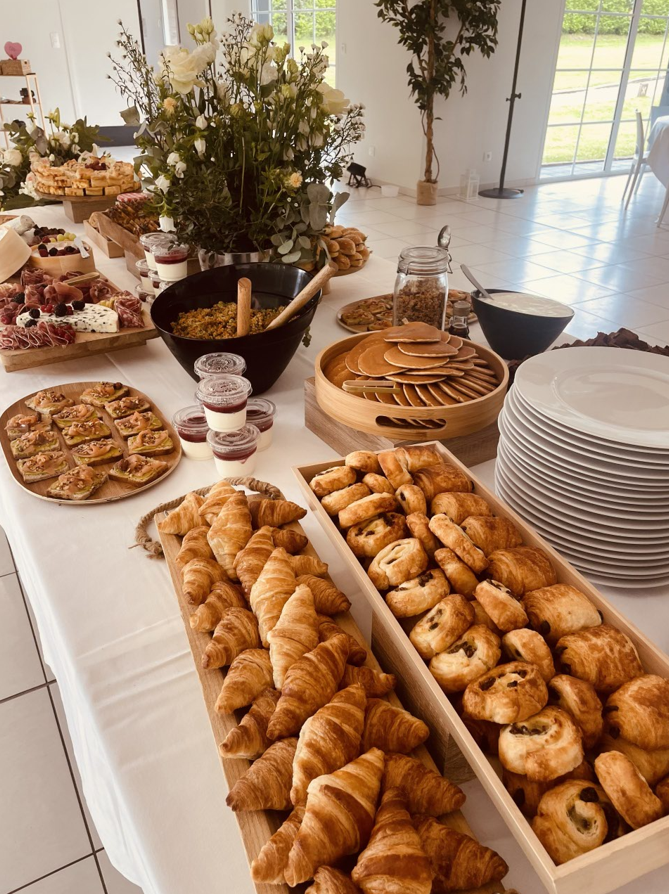 Buffet avec pâtisseries, fromages et fleurs, installé dans une pièce lumineuse avec de grandes fenêtres.