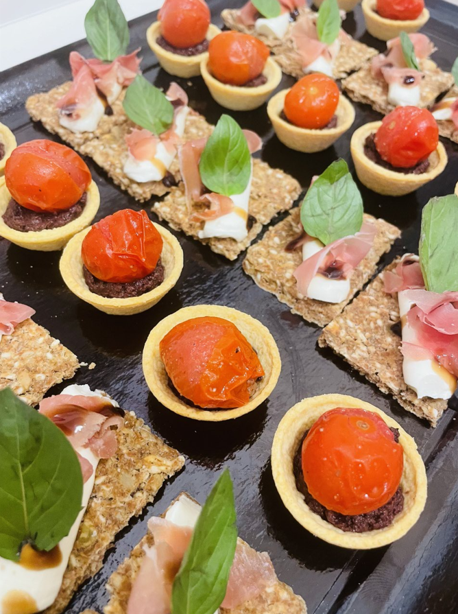 Plateau d'entrées : tartelettes aux tomates et craquelins avec mozzarella, prosciutto et basilic sur un plateau noir.