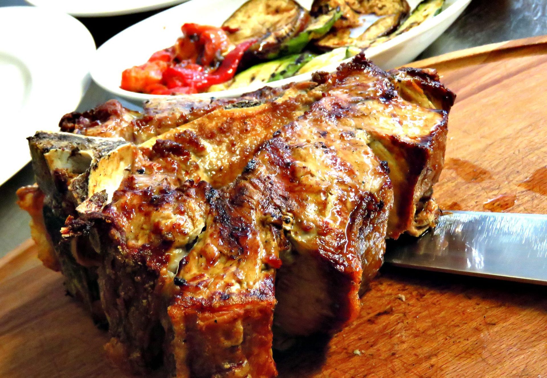 Costoletta di maiale arrosto su un tagliere di legno con verdure grigliate sullo sfondo.