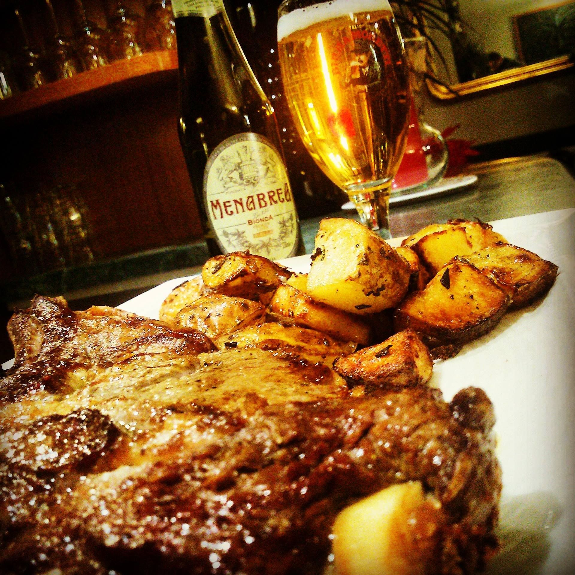 Bistecca e patate arrosto su un piatto bianco con birra e una bottiglia di vino sullo sfondo.