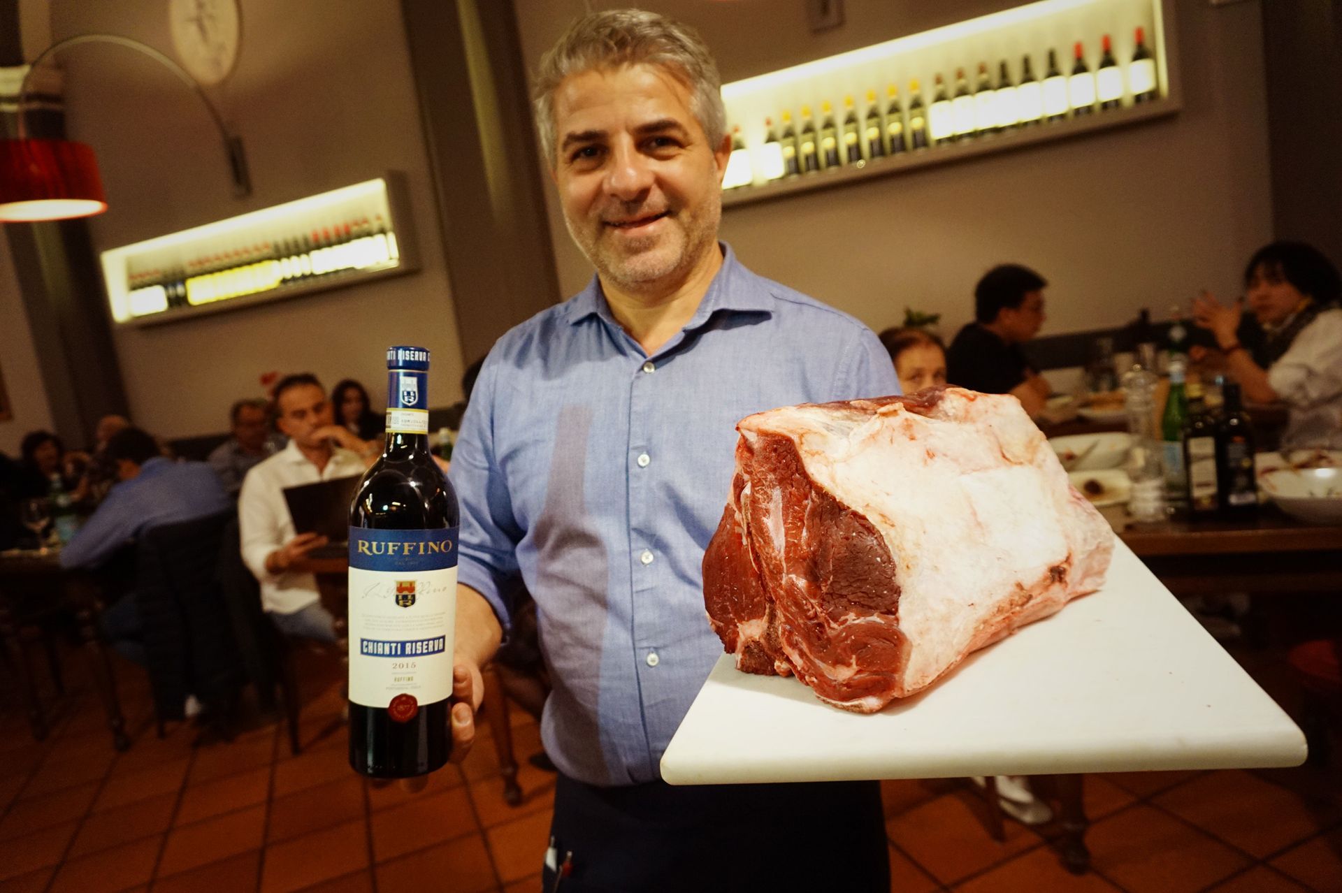 Uomo che tiene in mano una bottiglia di vino e una grossa bistecca cruda in un ristorante.
