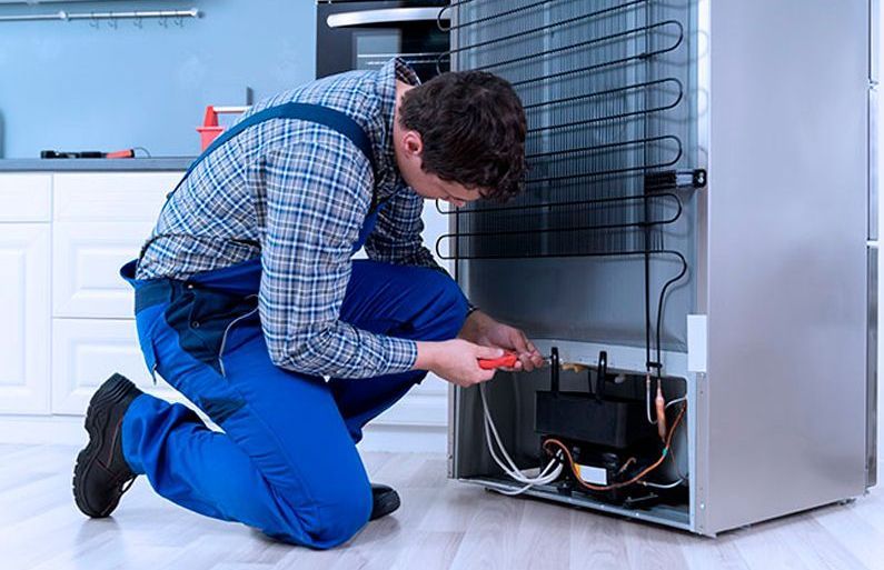 Service de heladeras arrodillado, usando un destornillador en la parte posterior de un refrigerador plateado en una cocina.