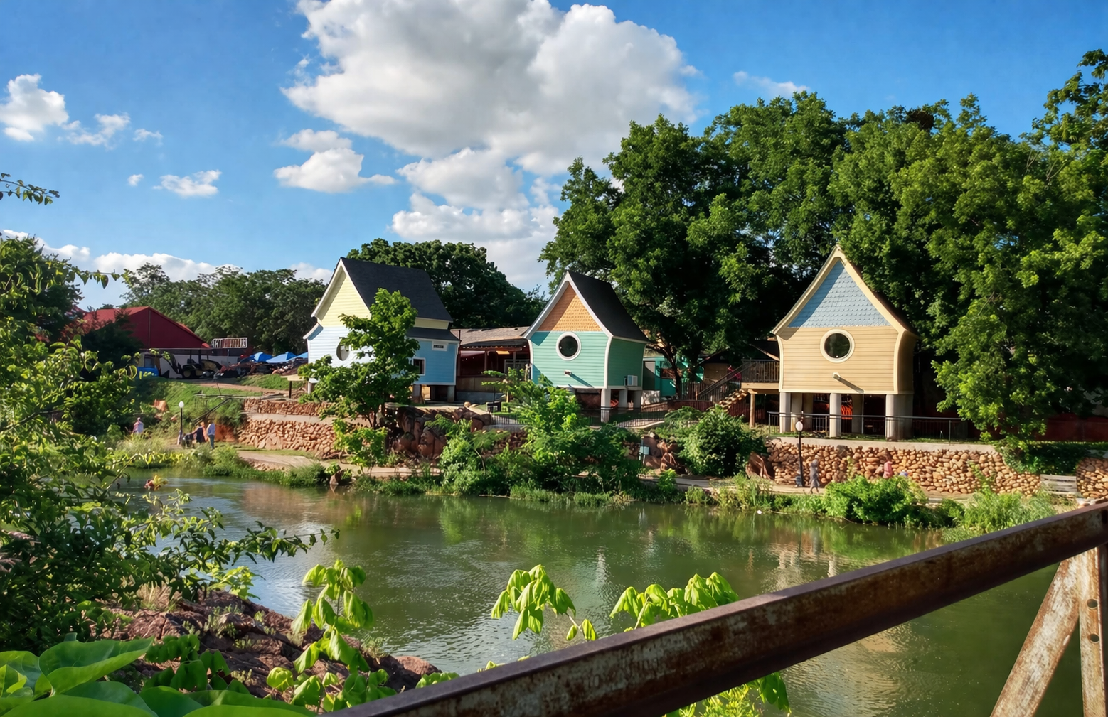 Scenic pond with flower beds and small cottages under a bright blue sky, viewed from a wooden bridge