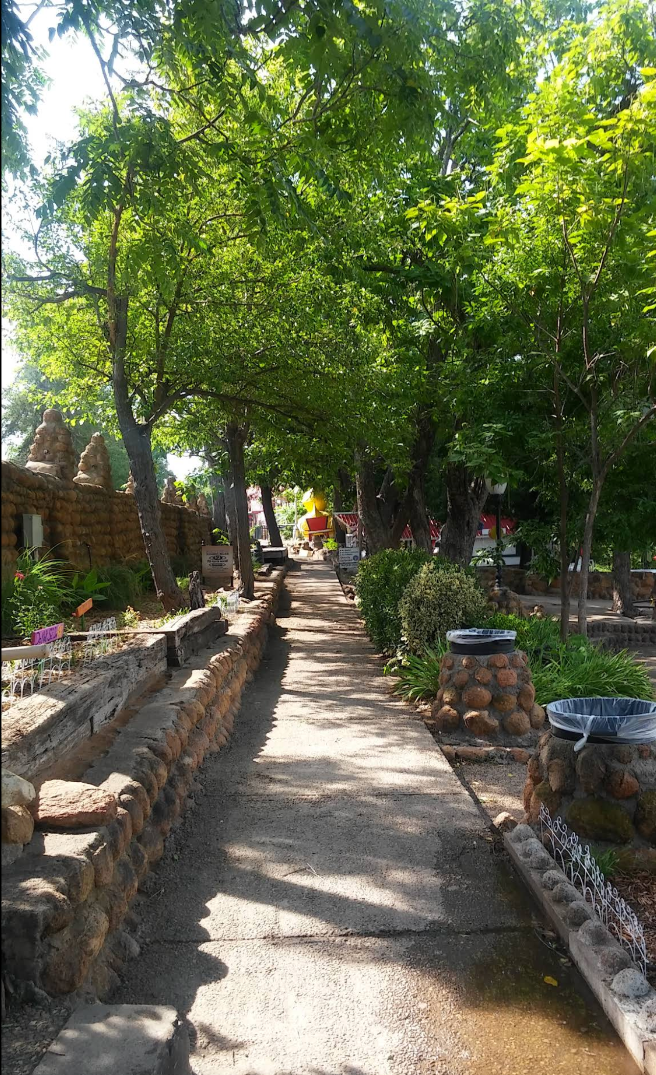 Shaded garden pathway lined with stone borders and lush green trees, with a few people in the distance