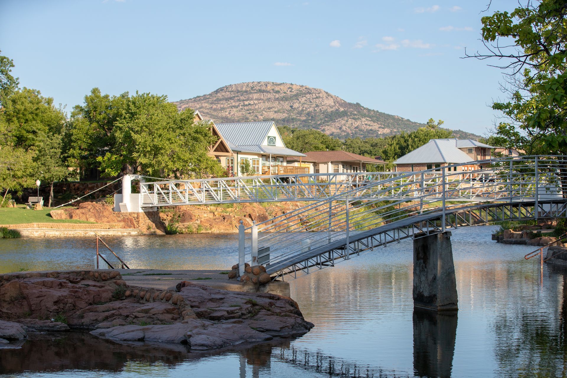 Suspension bridge over a calm river in a park, with trees, rocky banks, and a mountain under a blue sky